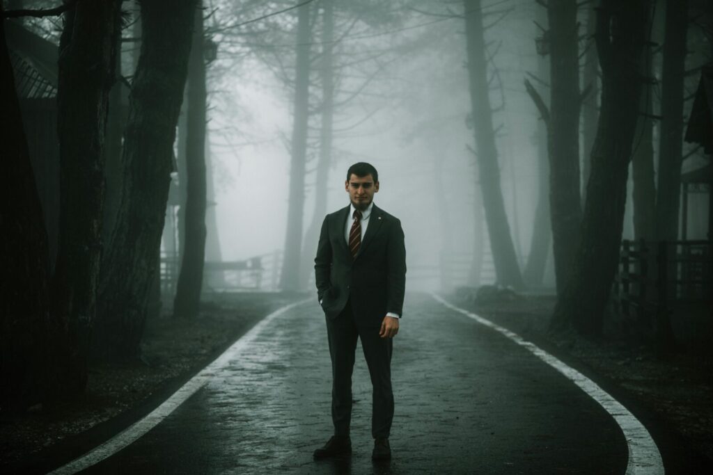 A solitary man in a suit stands on a foggy forest road, creating a moody atmosphere.