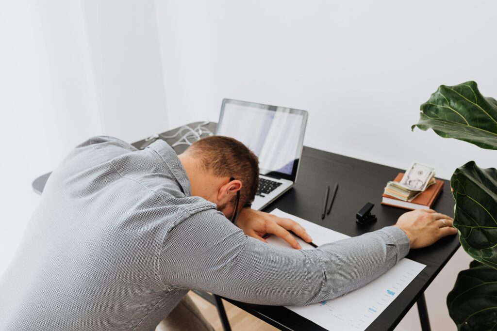 Tired man asleep at a desk with an open laptop and paperwork, indoors.